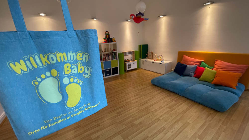 Das Bild zeigt einen Jutebeutel mit der Aufschrift "Willkommen Baby" vor dem Hintergrund eines Spielzimmers für Kleinkinder mit Spielküche und Kuschelecke