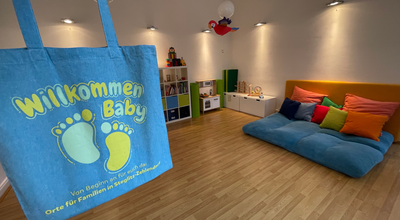 Das Bild zeigt einen Jutebeutel mit der Aufschrift "Willkommen Baby" vor dem Hintergrund eines Spielzimmers für Kleinkinder mit Spielküche und Kuschelecke