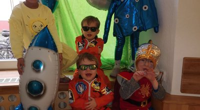 Kinder lachen und sind als Sonne, Alien und Astronauten verkleidet. Der Raum ist zum Thema Weltraum geschmückt.