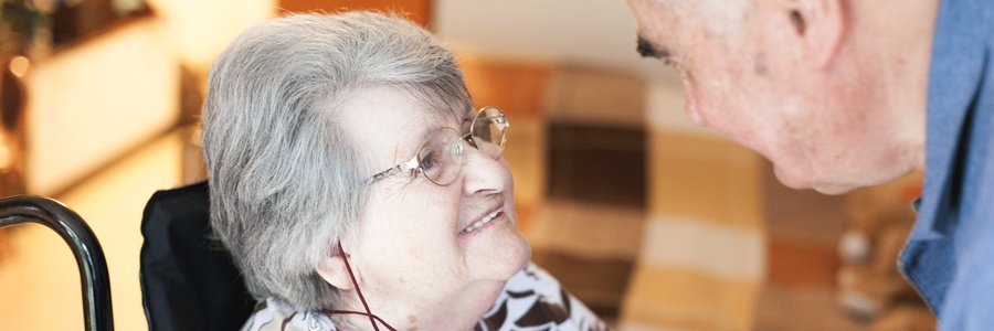 älterer Mann beugt sich liebevoll zu älterer Frau herunter, die im Rollstuhl sitz